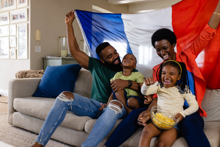 Happy african american parents with son and daughter with flag watching sport on tv at home. Childhood, family, togetherness, lifestyle, sport, entertainment and domestic life, unaltered.の写真素材