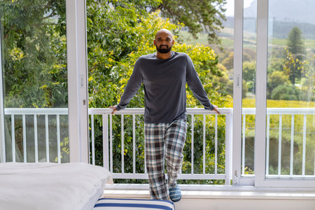 Portrait of happy biracial man in pyjamas standing at balcony in bedroom with treetops in background. Wellbeing, domestic life and lifestyle, unaltered.の写真素材
