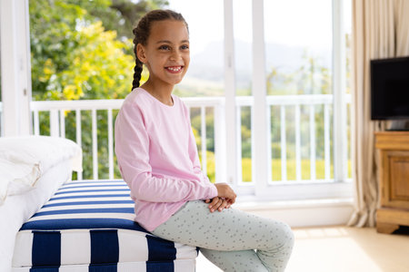 Happy biracial girl wearing pyjamas sitting in parents' bedroom in front of window to garden. Childhood, wellbeing, domestic life and lifestyle, unaltered.の写真素材