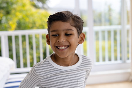 Happy biracial boy smiling in bedroom in front of window with view of garden. Childhood, wellbeing, domestic life and lifestyle, unaltered.の写真素材
