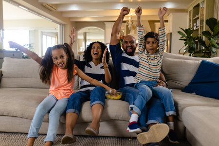 Happy biracial family sitting on couch with snacks, watching tv and cheering. Family, sport, entertainment, free time, togetherness, lifestyle and domestic life, unaltered.の写真素材