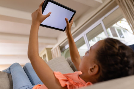 Happy biracial girl lying on couch using tablet with copy space on screen in living room. Childhood,...