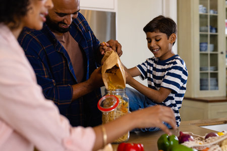 Happy biracial parents and son unpacking groceries and pouring pasta into storage jar in kitchen. Food, shopping, healthy lifestyle, ecology, family and domestic life, unaltered.の写真素材