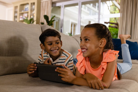 Happy biracial brother and sister lying on couch using tablet in living room. Childhood, free time, technology, communication, entertainment, lifestyle and domestic life unaltered.の写真素材