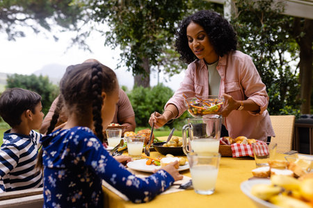 Happy biracial parents, son and daughter serving food and sitting at table for meal in garden. Summer, family, togetherness, food and lifestyle, unaltered.の写真素材