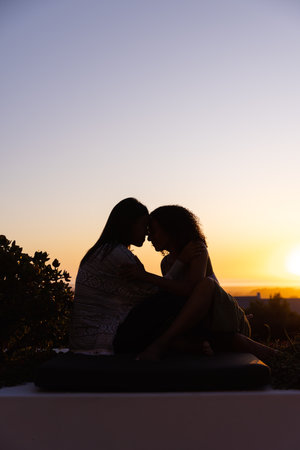 Biracial lesbian couple sitting and embracing in garden at sunset, copy space. Lifestyle, relationship, togetherness, nature and domestic life, unaltered.の写真素材