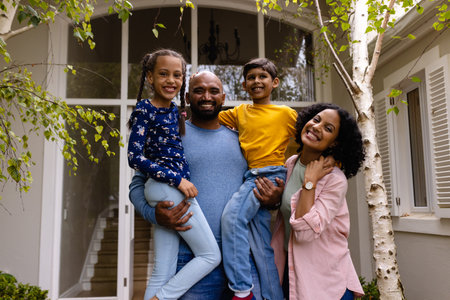 Portrait of happy biracial parents holding son and daughter, embracing in garden outside house. Family, love, togetherness, home, lifestyle and domestic life, unaltered.の写真素材