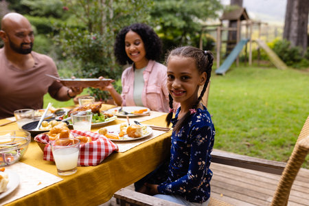 Portrait of happy biracial daughter at table having meal with parents in garden. Summer, family, togetherness, food and lifestyle, unaltered.の写真素材