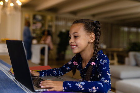 Happy biracial girl sitting at dining table using laptop in online class at home. Education, childhood, online learning, communication, lifestyle and domestic life, unaltered.の写真素材