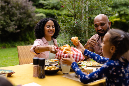 Happy biracial parents and daughter sitting at table having meal and talking in garden. Summer, family, togetherness, food and lifestyle, unaltered.の写真素材