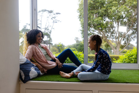 Happy biracial mother and daughter sitting by window to sunny garden and talking at home, copy space. Childhood, motherhood, relaxation, togetherness, lifestyle and domestic life, unaltered.の写真素材
