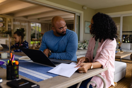 Biracial parents using laptop, looking at bills and talking at table with daughter in background. Finance, family, communication, lifestyle and domestic life, unaltered.の写真素材