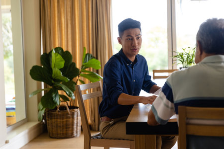 Happy diverse financial advisor and senior man discussing paperwork in dining room. Finance, retirement, domestic life and senior lifestyle, unaltered.の写真素材