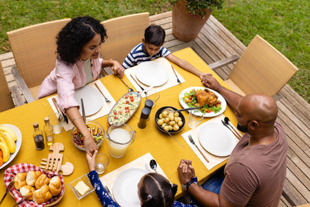 Happy biracial parents, son and daughter praying before meal, holding hands at table in garden. Summer, family, togetherness, religion, food and lifestyle, unaltered.の写真素材