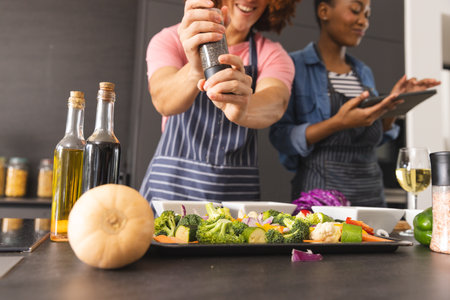 Happy diverse couple preparing meal, using tablet and seasoning vegetables in kitchen. Food,...