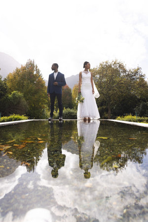 Happy african american groom and bride holding bouquet reflected in pond in sunny garden, copy space. Marriage, romance, summer, tradition, ceremony, nature and lifestyle, unaltered.の写真素材