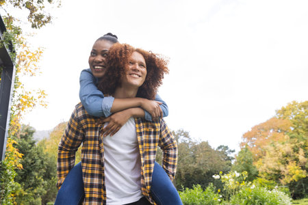 Happy diverse couple having fun piggybacking in sunny garden, copy space. Togetherness, relationship, free time, lifestyle, fun and romance, unaltered.の写真素材