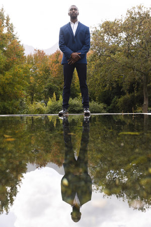 Thoughtful african american groom reflected in pond standing in sunny garden, copy space. Marriage, romance, summer, tradition, ceremony, nature and lifestyle, unaltered.の写真素材