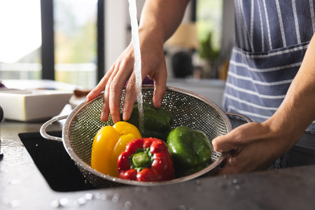 Midsection of biracial man in apron washing peppers in kitchen sink. Food, cooking, healthy...