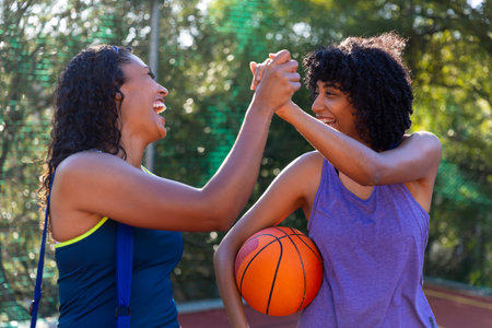 Two happy biracial female friends holding hands and basketball at basketball court. Sport, activity, friendship and lifestyle, unaltered.の写真素材