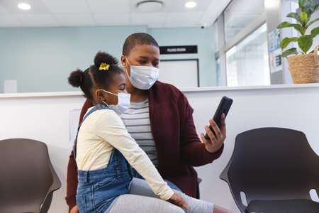 African american mother and daughter wearing face masks using smartphone in waiting room at...