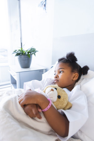 Sad african american girl patient lying on bed with teddy bear in patient room at hospital. Hospital, childhood, medicine and healthcare, unaltered.の写真素材
