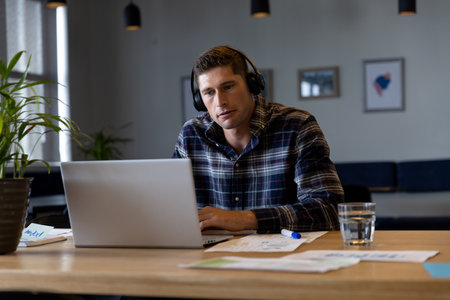 Caucasian casual businessman making video call using using headphones and laptop at desk. Casual office, communication, business and work, unaltered.の写真素材