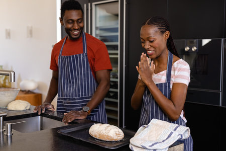 Happy african american couple in aprons enjoying baked bread in kitchen. Food, cooking, lifestyle,...