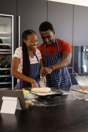 Happy african american couple in aprons preparing bread dough in kitchen. Food, cooking, lifestyle,...