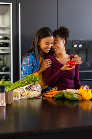 Happy biracial lesbian couple embracing and unpacking groceries in kitchen. Shopping, food, healthy lifestyle, lifestyle, relationship, togetherness and domestic life, unaltered.の写真素材
