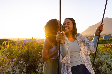 Happy biracial lesbian couple kissing and swinging on swing in sunny garden. Lifestyle,...