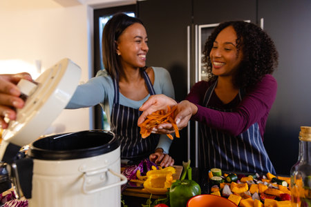 Happy biracial lesbian couple composting vegetable waste in kitchen. Cooking, food, healthy...