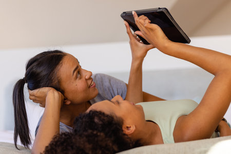 Happy biracial lesbian couple using tablet on bed in bedroom. Relaxation, lifestyle, relationship,...
