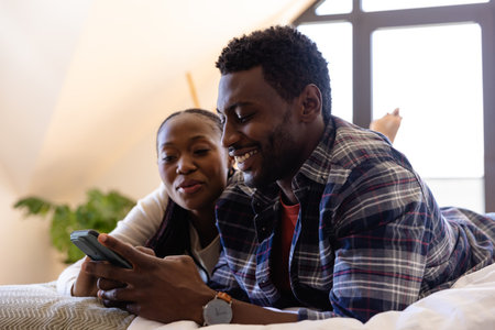 Happy african american couple using smartphone lying on bed in bedroom. Relaxation, lifestyle,...