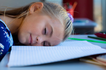 Tired caucasian schoolgirl sitting at desk and lying on books in classroom at elementary school....