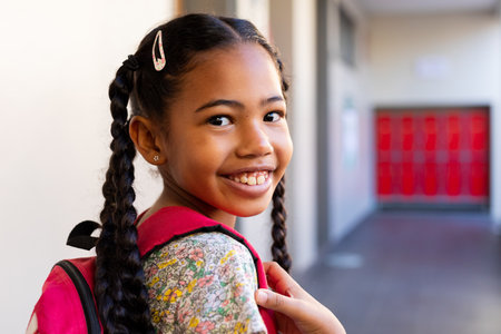 Portrait of happy biracial schoolgirl with school bag in corridor at elementary school. Education, childhood, development, learning and school, unaltered.の写真素材