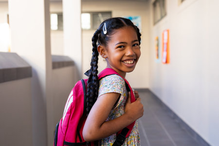 Portrait of happy biracial schoolgirl with school bag smiling in corridor at elementary school. Education, childhood, development, learning and school, unaltered.の写真素材