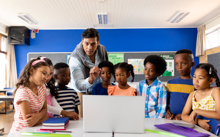 Biracial male teacher using laptop teaching children in elementary school class. School, learning, childhood, communication, teaching and education, unaltered.の写真素材