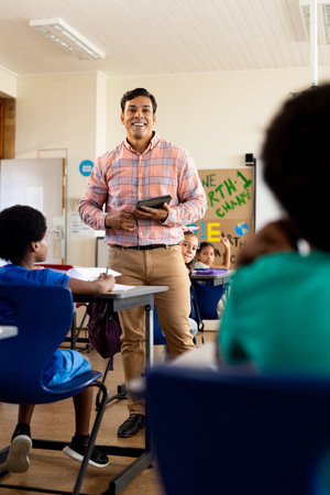 Diverse male teacher with tablet and elementary schoolchildren in class at elementary school....