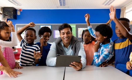 Diverse male teacher with tablet and elementary schoolchildren raising hands in class, slow motion....