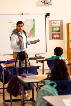 Diverse male teacher using tablet teaching with children at desks in elementary school class....