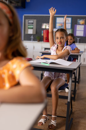 Diverse elementary schoolchildren raising hands at desks in class. School, learning, childhood, communication and education, unaltered.の写真素材