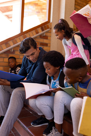 Happy diverse male teacher and children with books sitting on stairs in class at elementary school....
