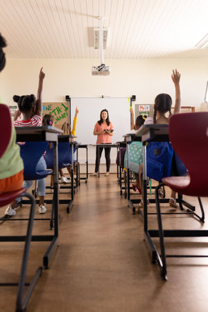 Diverse female teacher with tablet and elementary schoolchildren raising hands in class. School,...