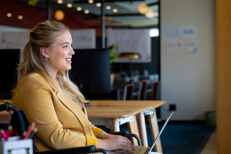 Happy plus size caucasian casual businesswoman in wheelchair using laptop in office. Casual office,...