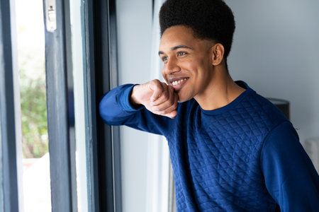 Happy biracial man standing in bedroom looking out of window, smiling. Free time, lifestyle, wellbeing and domestic life, unaltered.の写真素材