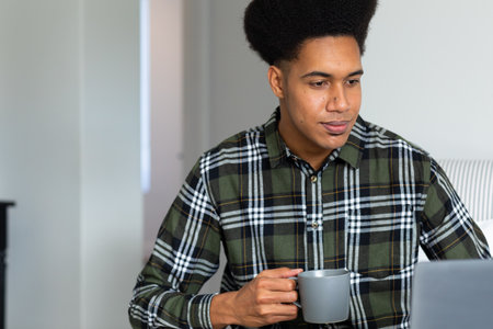 Happy biracial man sitting at home holding cup of coffee and using laptop. Free time, communication, lifestyle and domestic life, unaltered.の写真素材