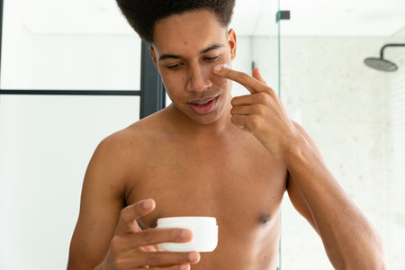 Biracial man looking at face cream and applying to face in sunny bathroom. Self care, skin care, wellbeing and lifestyle, unaltered.の写真素材