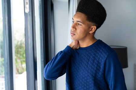 Thoughtful biracial man standing in bedroom looking out of window. Free time, lifestyle and domestic life, unaltered.の写真素材