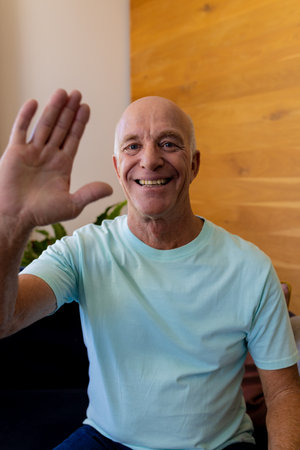 Happy senior caucasian man making video call smiling and waving at home. Senior lifestyle, communication, technology and domestic life, unaltered.の写真素材
